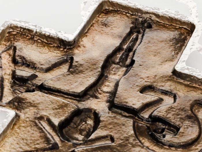 Nahaufnahme der Oberfläche: das Relief hebt sich in dunklen, weichen Graten aus dem Grund, die Gusshaut ist als feine Körnung zu sehen