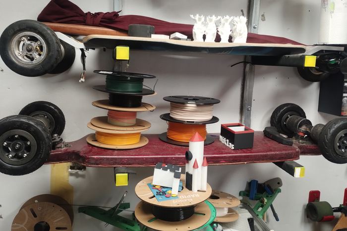Ein Werkstattregal mit Filamentrollen in vielen Farben, darüber Skateboard-Decks und Rollen