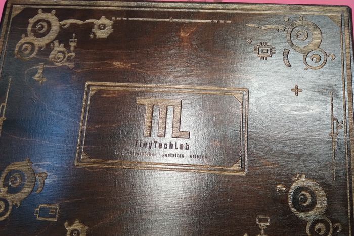 Eine große dunkle Holzplatte mit eingraviertem TinyTechLab-Zeichen und Zahnradmustern an den Ecken