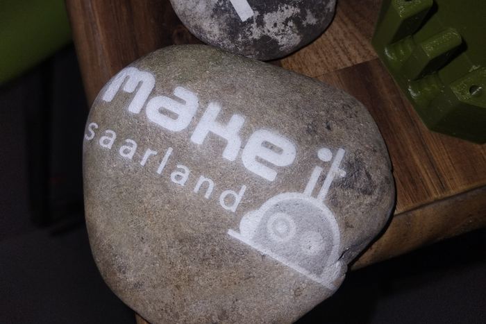 Zwei glatte Flusssteine mit eingravierten Schriftzügen, einer trägt das TinyTechLab-Zeichen, der andere „make it saarland“
